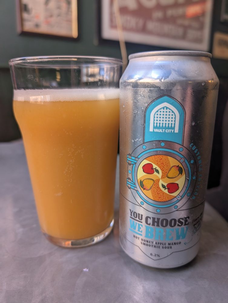 A pint glass filled with hazy orange beer sits next to a silver can of Vault City Brewing’s “You Choose We Brew” Hot Honey Apple Mango Smoothie Sour, 6.2% ABV. The can features an illustrated porthole design with apples and mangoes inside.

