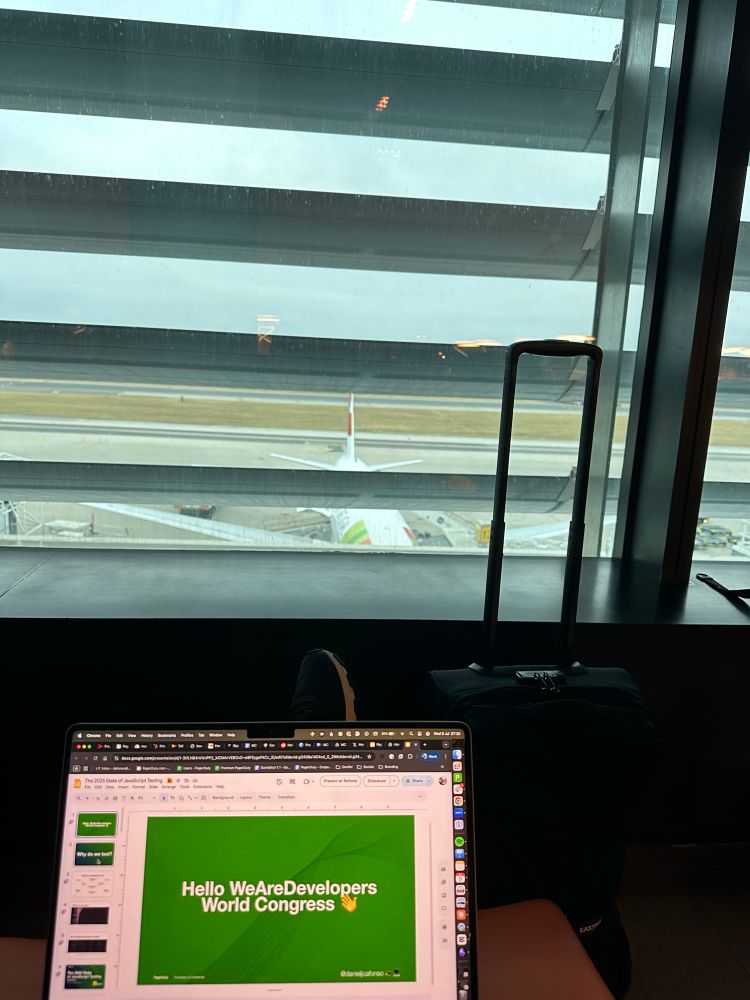 A person sits at an airport terminal with their legs stretched out, facing a large window overlooking the runway. In the foreground, an open laptop displays a presentation slide that reads “Hello WeAreDevelopers World Congress 👋” on a green background. A black suitcase stands upright next to the person, and an airplane is visible outside on the tarmac through the window’s horizontal blinds. The scene suggests the person is preparing for or traveling to a tech conference.