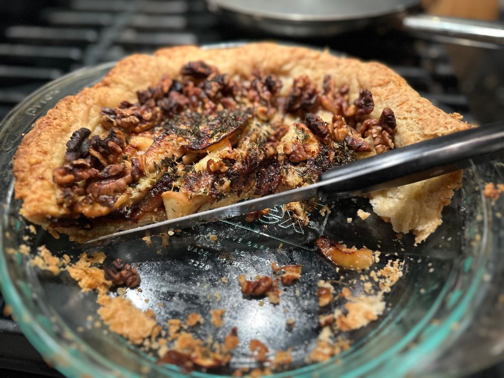 an ios portrait mode photo of a savory tart in a pyrex dish, with a thick brown crust and half of the tart missing. thyme leaves, walnuts, apples and onions are visible. a black and silver knife lies on top of the tart remains. the dish sits on a black stovetop.