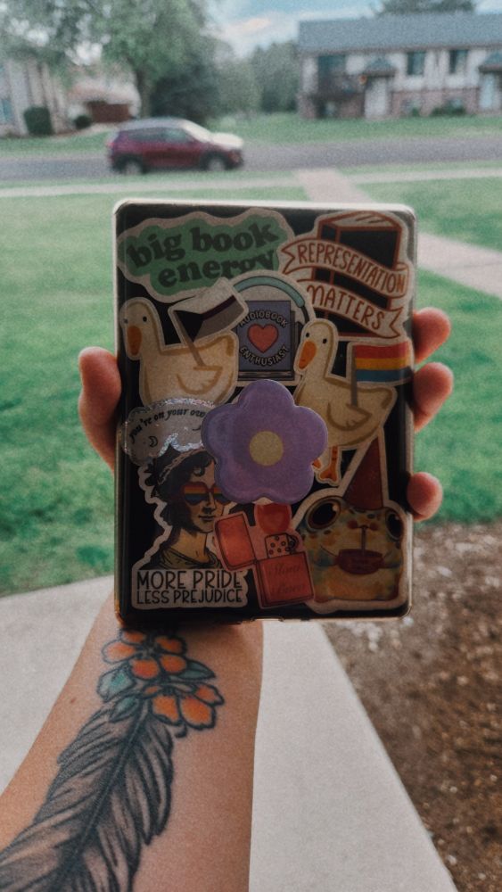 An arm holding out a Kindle to display the stickers on the back. A green sticker that says "big book energy", a black book stickers that says "representation matters", a goose holding a demisexual flag, a book with headphones around it that reads "audiobook enthusiast", another goose sticker this with the pansexual flag, a Victorian woman with rainbow heart sunglasses with the words "more pride less prejuidice" under it, a pink lighter that says slow burn and lasts a plump frog with a dunce cap on drinking a cup of "dumb juice"