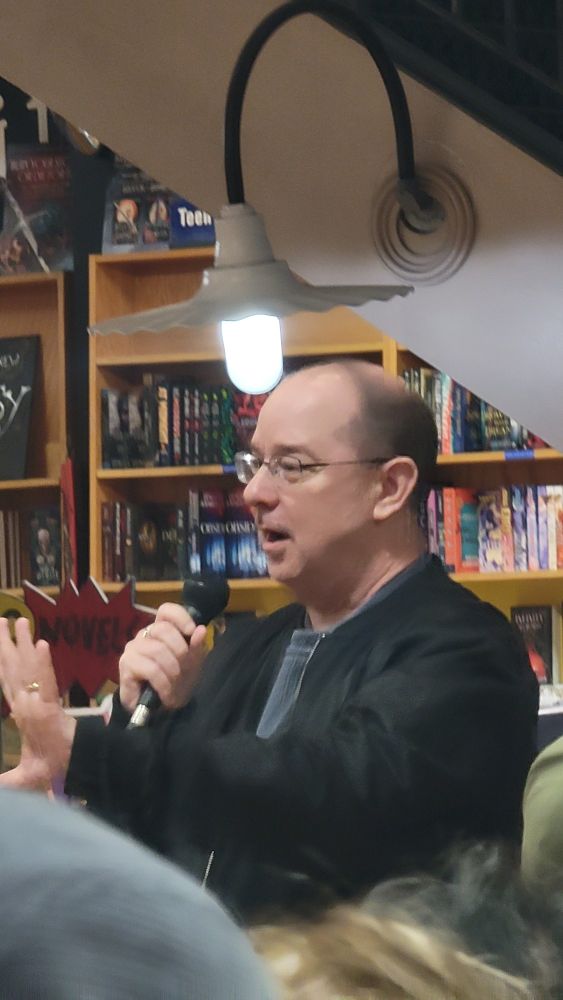 Author John Scalzi, a middle aged white man standing in a crowded bookstore speaking into a microphone. 