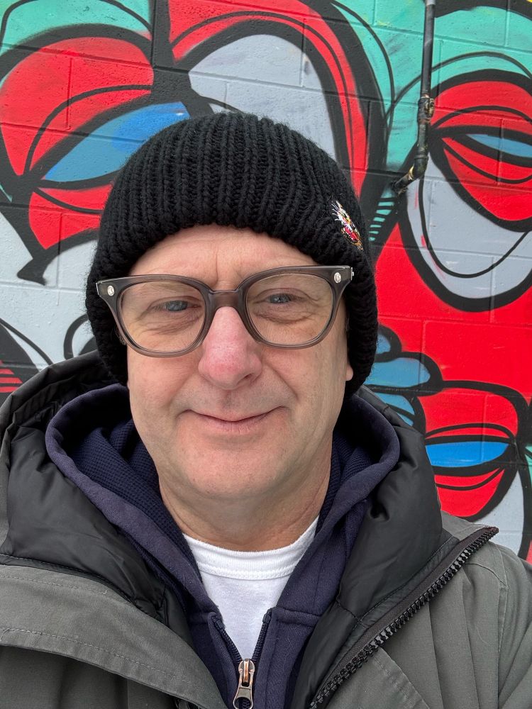 Selfie of an elder Canadian man with vintage glasses, wearing a toque, hoodie, and winter jacket (hood down), with a graffiti wall in the background (it’s a nice piece by another local artist, not the man pictured)