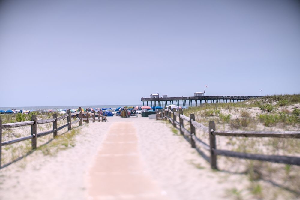 A soft-focus view down a sandy beach walkway framed by wooden fences, leading to a vibrant cluster of umbrellas and beachgoers near the ocean. A wooden pier extends from the right into the sea under a clear, blue sky. The center of the image is sharply focused while the edges blur gently, emphasizing the boardwalk and ocean ahead.