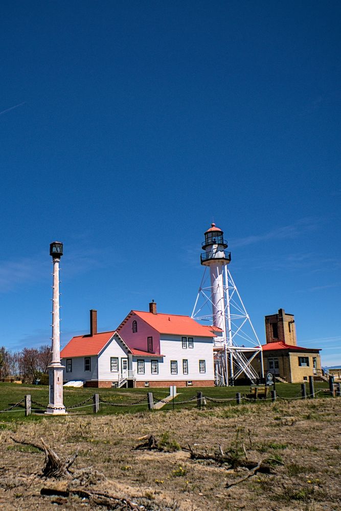 Historic lighthouse and keeper’s quarters – A white-painted Whitefish Point Light Tower, with a bright red-roofed keeper’s house and fog station, standing on grassy terrain against a deep blue sky.