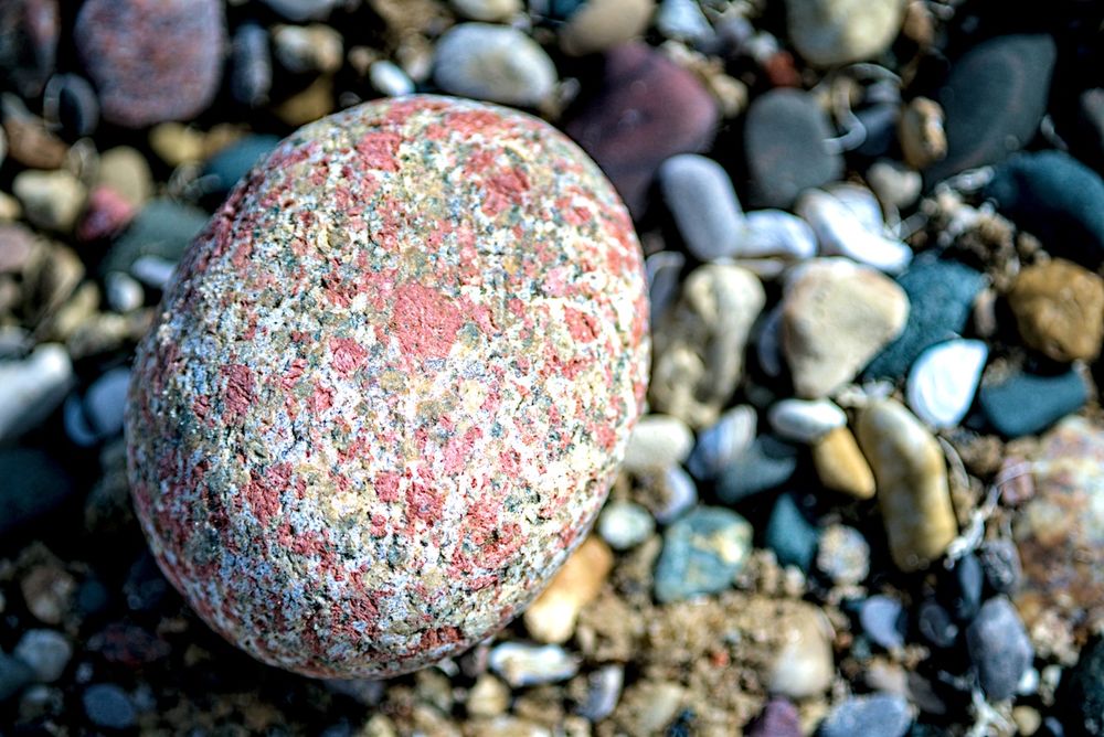 Colorful beach stone – A close-up of a large, smooth, multicolored rock with red, white, and gray speckles resting among smaller pebbles on a beach.
