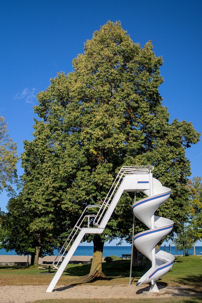 A white spiral slide stands in a park in Rogers City, MI, surrounded by lush green trees. The slide has red and blue accent stripes and is set on a sandy play area. Behind it, picnic tables and benches are scattered beneath the trees, with Lake Huron's blue waters and a sandy beach visible in the background. The scene is bathed in bright sunlight under a clear blue sky.