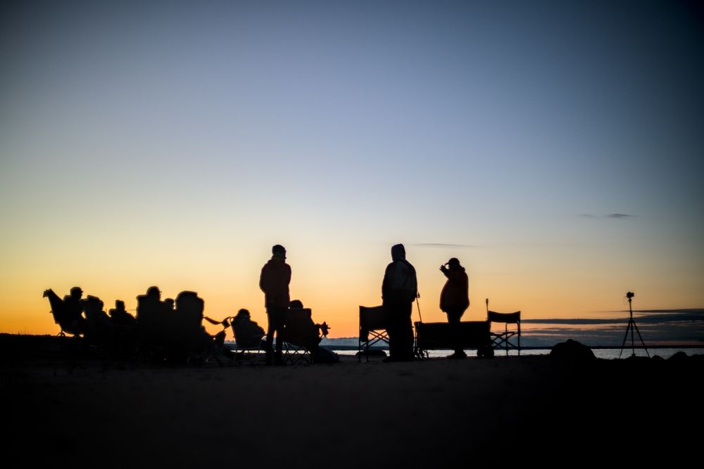 Silhouetted figures gather in quiet conversation along the sandy beach at dusk. The sky glows softly from the fading sun, casting cool light over the scene. Folded chairs and a single tripod are silhouetted in the stillness of evening.

Camera & Processing Details:
Photos taken with a Sony a7RII using a Pentax Super-Takumar 50mm F1.4 vintage camera lens. Images were processed in Darktable.
