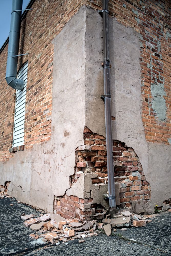 Crumbling brick corner with drainpipe – The base corner of a brick building where plaster has fallen away, revealing red bricks and a rusty downspout, with scattered debris on the pavement.