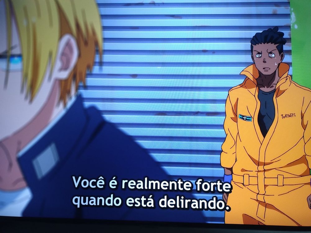 Cena do anime "Fire Force" (Enen no Shouboutai) em que um personagem negro de nome Ogum, ao fundo, fala para Artur, loiro, que está em sua frente a frase "você é realmente forte quando está delirando"