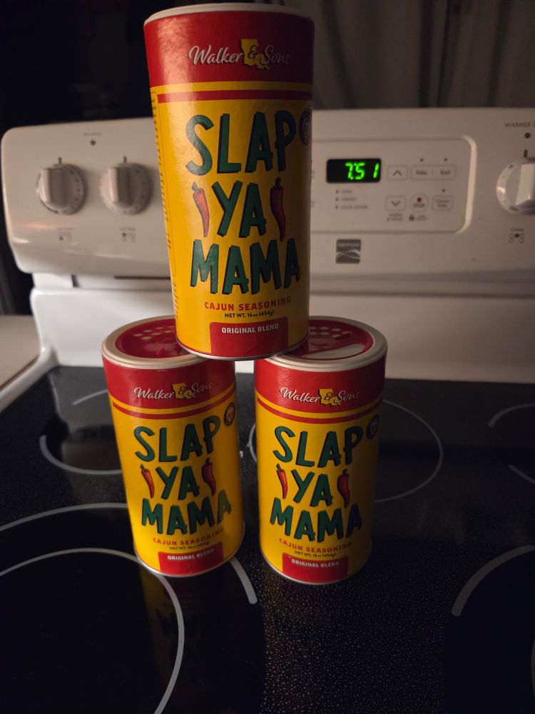 Stacked on the black glass surface of a white stove are three massive yellow Slap ya mama cajun spices jar.