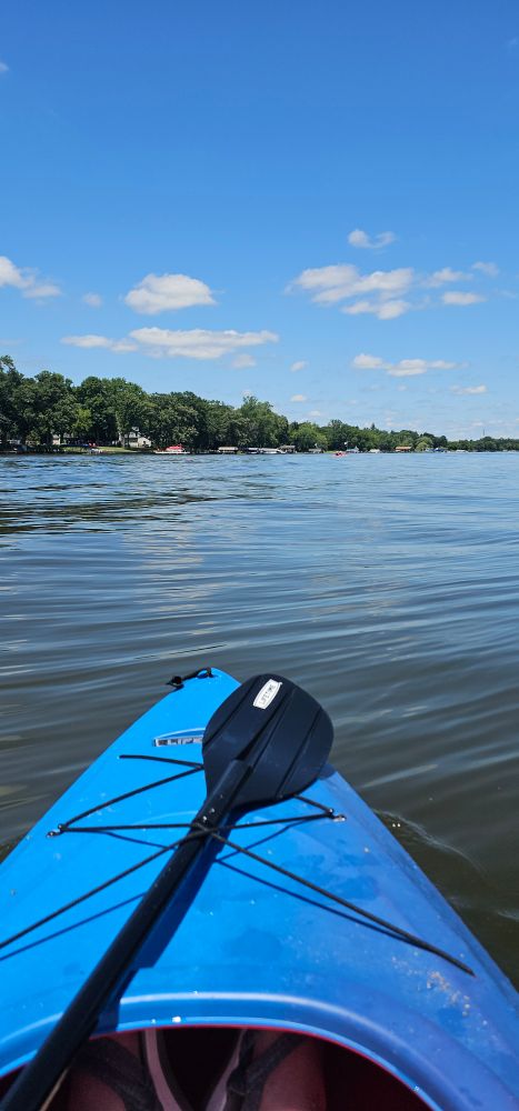 The nose of a blue plastic kayak with a black paddle at the bottom of the image, floating on a river bordered by large trees and houses. The river reflects the beautiful blue sky dotted with tiny clouds. Off in the distance, my friends are in an orange kayak you can barely see. 