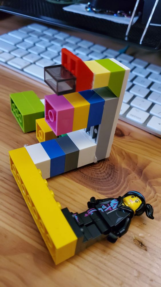 A contraption made of multi-colored Lego blocks with a minifig of Wyld Style from The Lego Movie on one end, laying on a wooden table in front of a Mac keyboard.
