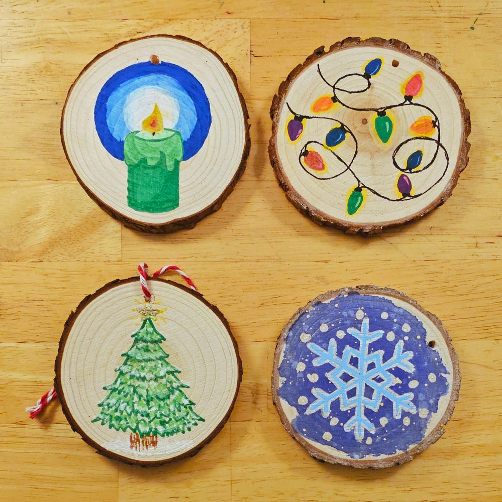 Four wood-slice ornaments with Christmas-themed illustrations. From top left, a green candle with a blue halo around the flame, a string of glowing Christmas lights, a silver and blue snowflake, and a snowy pine tree 