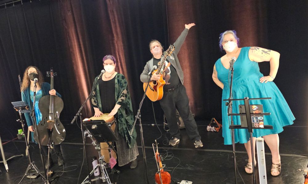 The Vixy & Tony band on stage! From left to right: Betsy Tinney, cellist, a small pale woman in blues holding a carbon fiber cello; Vixy Dockrey, lead singer and drummer, a pale woman wearing a black shirt and green kimono jacket, standing in front of a djembe drum; Tony Fabris, guitarist, a white man wearing black and grey and holding a guitar; and me, violinist, a white woman wearing a teal dress. The band is all wearing masks because this is a Clean Air Events show that protects the audience and performers alike. 