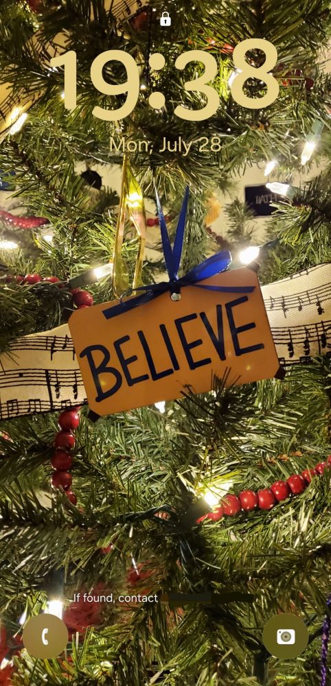 A Christmas tree ornament of the "BELIEVE" sign from Ted Lasso (dark blue writing on a yellow rectangle), hanging on a tree with lights, a red wooden cranberry garland and a ribbon garland with sheet music on it. The time is 19:28, the date is Mon, July 28.