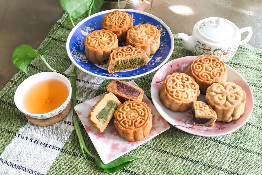 Photo of a spread of mooncakes with various designs.  Some of them are cut open to reveal pistachio, strawberry, matcha, and red bean fillings.  The spread also includes a small teapot and cup of tea.