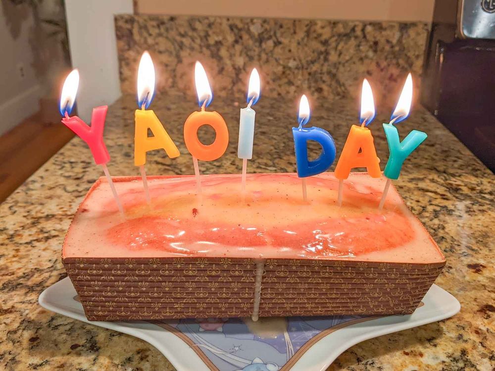 Photo of a cake with letter-shaped candles spelling out yaoi day.  The candles are on fire