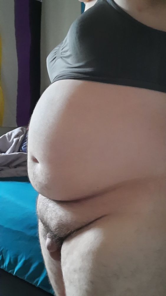 A girl in a dark bra poses with her tummy and clit exposed for Tummy Tuesday. The nonbinary flag hangs in the background.