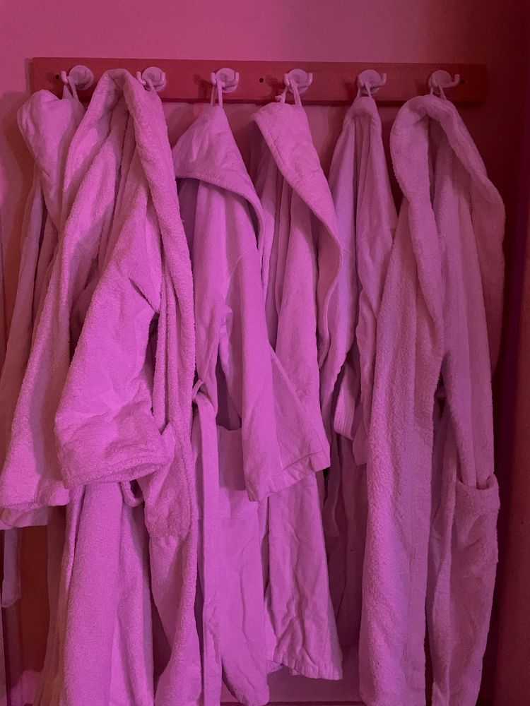 Einige Bademäntel nebeneinander auf Haken, rosa beleuchtet. 