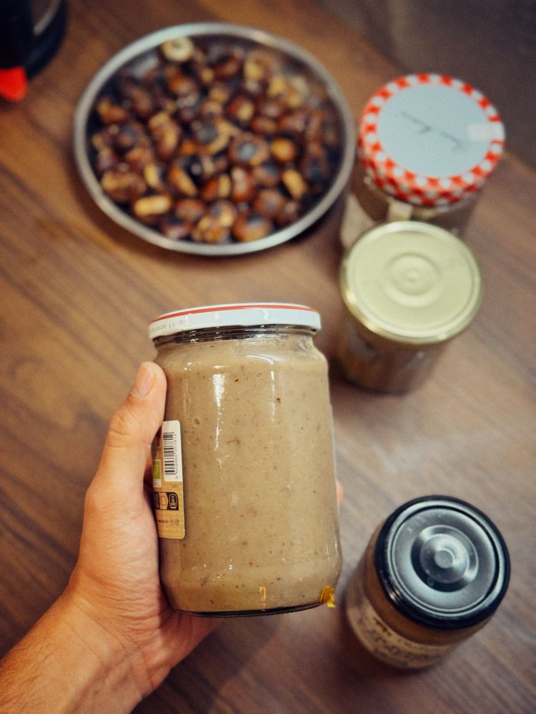 Photo d’un pot de crème de marron tenu dans ma main gauche. Au second plan, 3 autres pots et des châtaignes grillés 