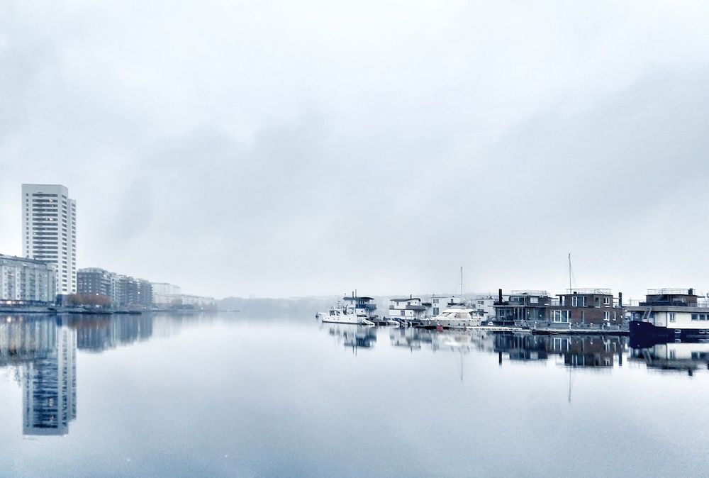 En grå disig dag. Sjön ligger spegelblank. till höger vid horisontlinjen ligger bostadspråmar och till vänster bostadshus. Allt speglas i vattnet. 