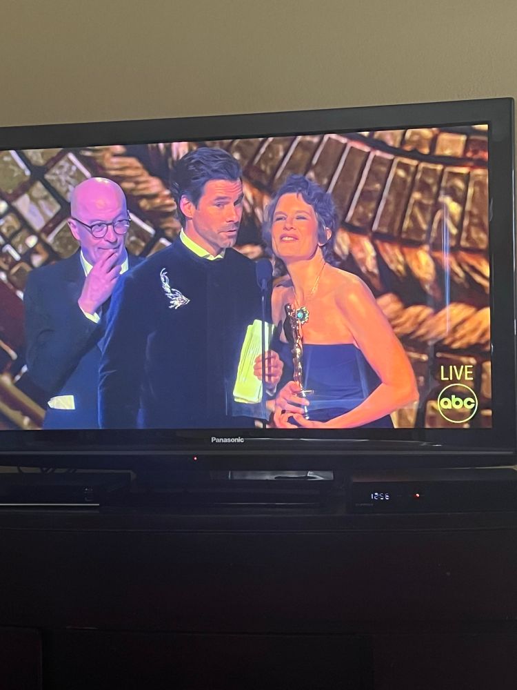 A man and woman accept their Oscars, while another man hovers behind, with no access to the mic. 