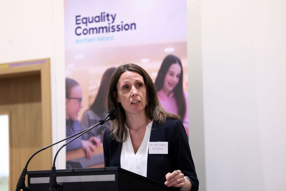 Dr Eve Hepburn speaks at a podium with a microphone, with an Equality Commission for Northern Ireland banner in the background
