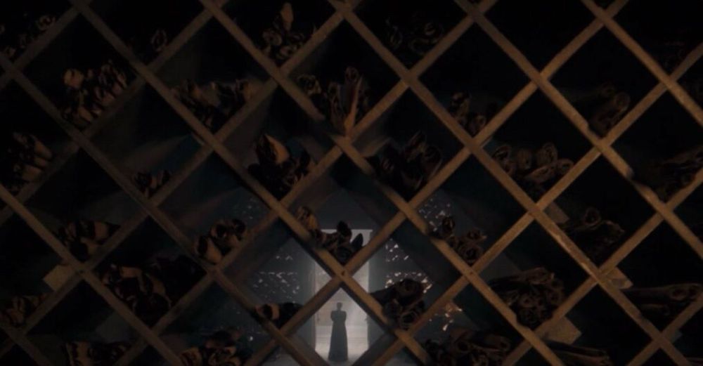 one of the last shots of season 2 of house of the dragon. rhaenyra targaryen is tiny in the bottom center of the frame, distant, scaled and positioned the same as alicent in the previous shot. positioned between her and the camera is the scroll shelf with the lattice shape, and she is framed perfectly by one of the openings in it. the lattice shaped shelf is dark and overbearing, appearing to trap her, and only the doorway rhaenyra stands in in the distance offers a tiny sliver of light, rendering her in silhouette. she is trapped.
