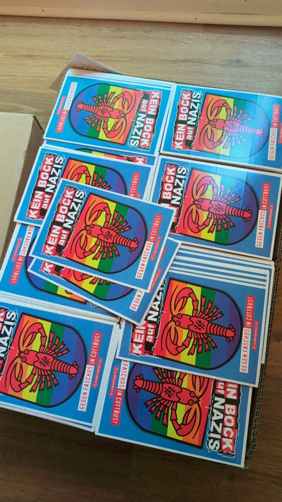 Eine Kiste mit Karten auf deinen steht "Kein Bock auf Nazis" Dazu ein Krebs auf einem Regenbogenfarbenen Wappen.