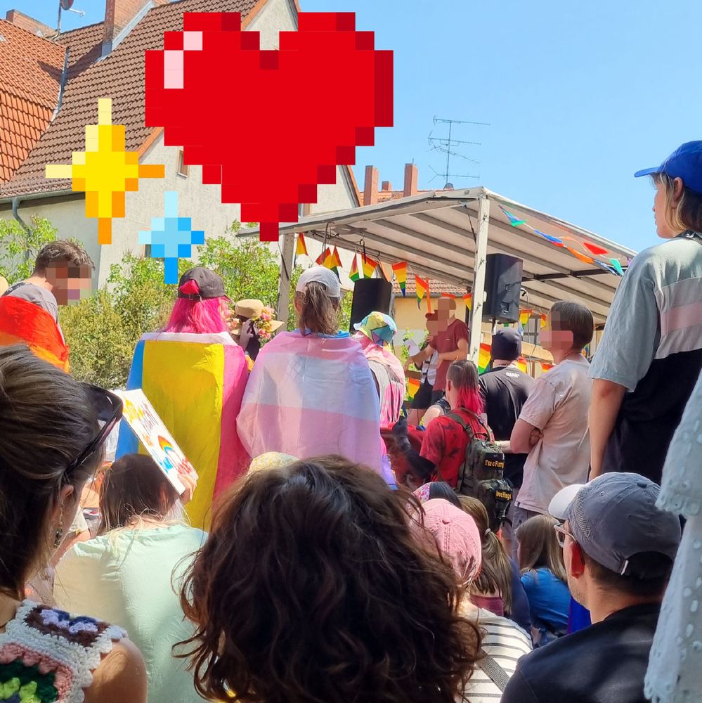 Foto von Pride-Demoteilnehmenden von hinten, mit Blick in Richtung eines Lautsprecherwagens. An Menschen und Wagen hängen verschiedene Pride-Flaggen. Im Hintergrund steht ein graurotes Einfamilienhaus, darüber sind herz und sternförmige Sticker 