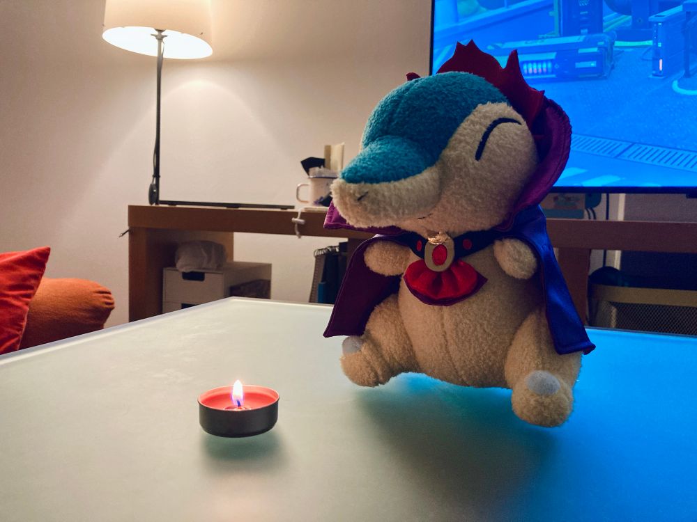 My cute dracula cyndaquil plushie and a candle on a table with my living room as background (my TV, lamp, laptop…)