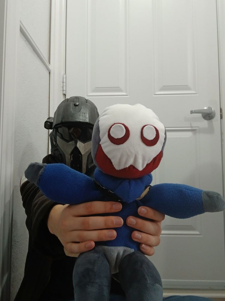 A person wearing a helldiver helmet holds a plush automaton wearing a blue sweater towards the camera