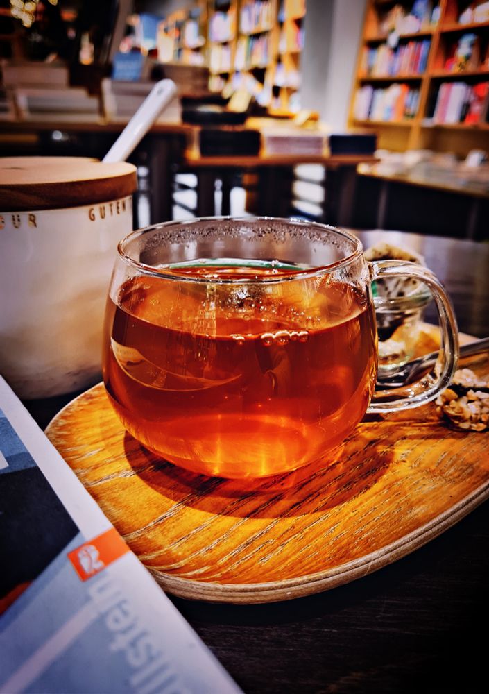 ein kleines Holztablett, darauf eine Glastasse mit Tee, daneben liegt ein Buch, im Hintergrund Regale und Tische eines Buchladens.