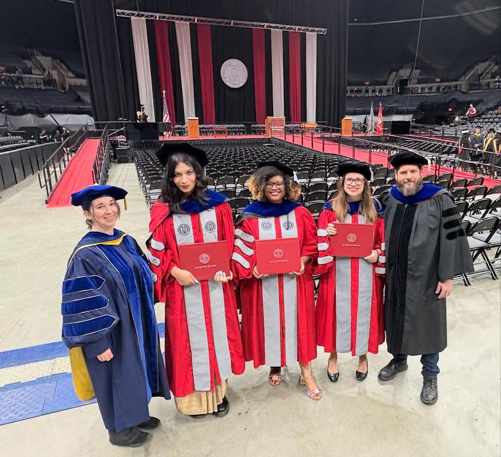 Image from 2025 summer graduation at Ohio State with 5 people in PhD regalia in front of empty seats and a stage with the OSU seal. L to R: Laura Lopez, Joy Bhattacharyya, Caprice Phillips, Alison Duck, Scott Gaudi.
