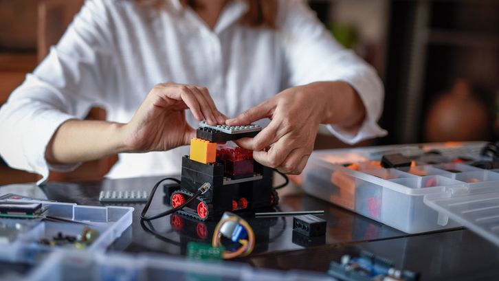 A student in a white shirt assembling a small wheeled robotic vehicle using colorful plastic bricks and electronic components. She is carefully placing a flat LEGO-like piece on top, surrounded by open storage boxes filled with additional parts.