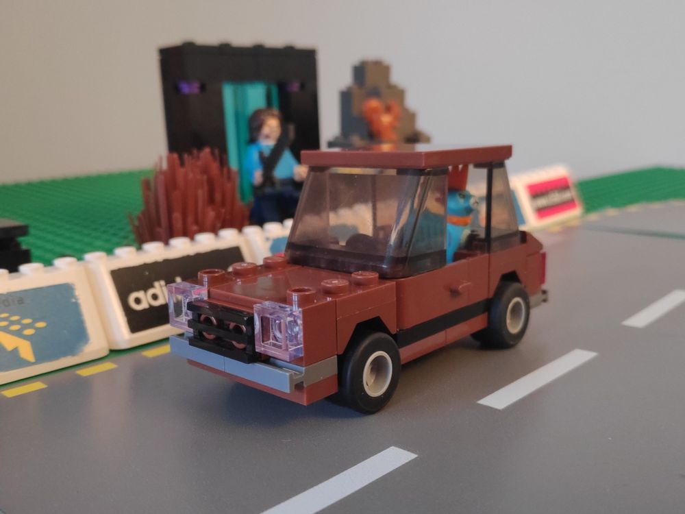 Lego vignette of a 4-wide old-looking reddish-brown car with a cat in the backseat on a road edged by banner ads. An out-of-focus Jack Black exits an Earth Portal in the background.
