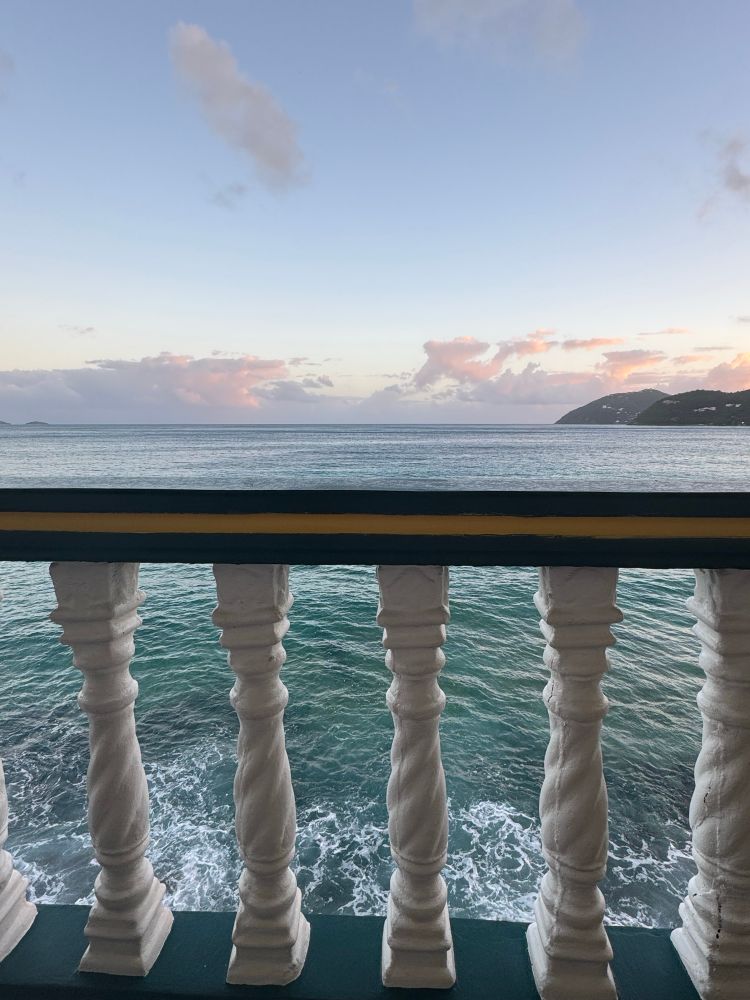 View from a balcony: The ocean crashes on shore through the painted wooden railing with white vertical spindles and yellow and green horizontal top and bottoms . Sun rise gives way to red and purple clouds at the horizon of the water and a pale blue above that.   Land pokes into the frame on the far right. 