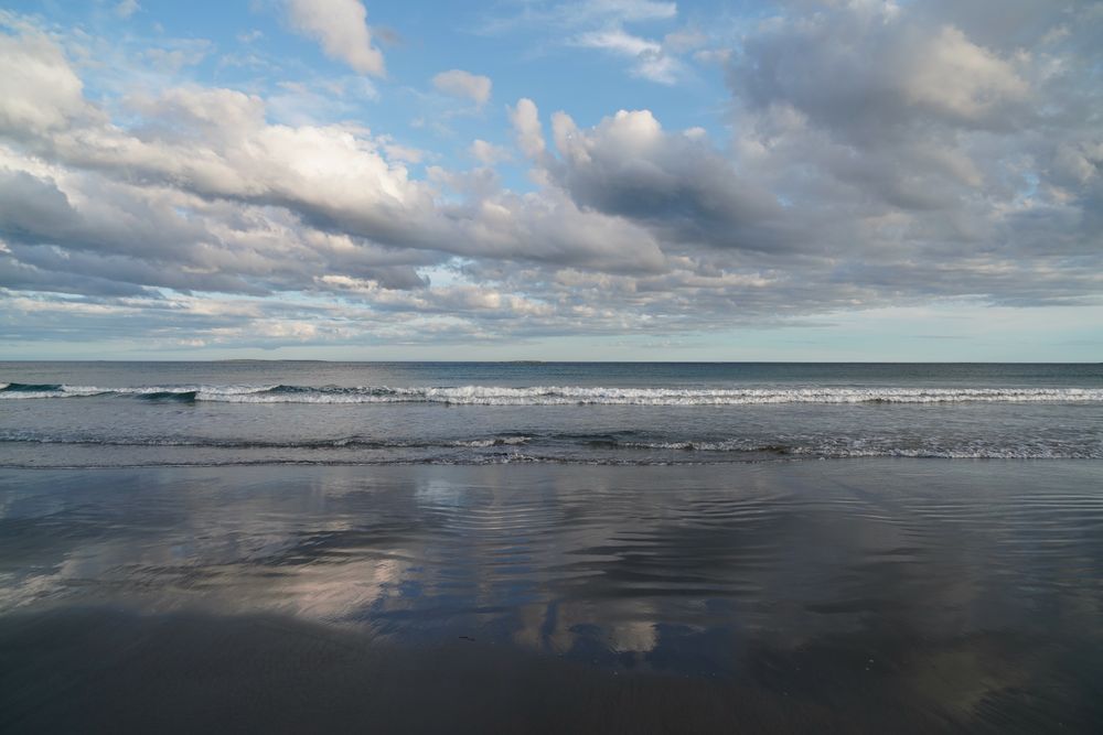 Beach …grey-white clouds in the blue sky in the top half of the frame reflect on the smooth wet sand on the bottom of the frame. Waves lap the shore through the middle of the frame 