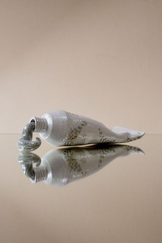 Salt fired porcelain nerikomi toothpaste tube. It is white with some pastel pattern and it’s squeezing out a bit of striped toothpaste. It sits on a mirror on a beige background 