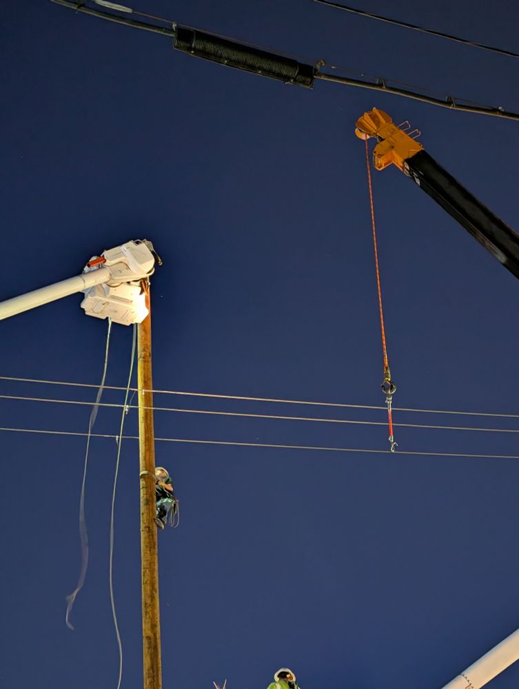 indigo night sky, as a lineman climbs with no visible harness up to the top of the new pole, cranes in the foreground waiting to help him do his work