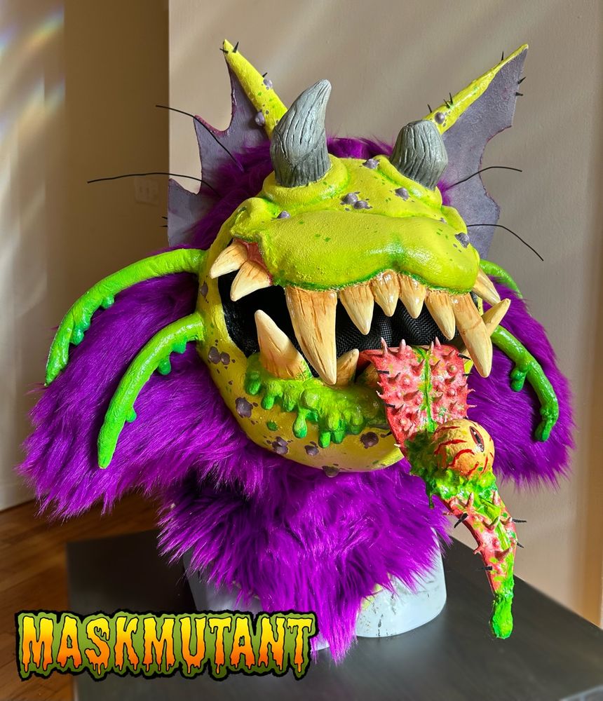 Costume head of a neon green alien cat with purple fur, puking slime. Its eyeball is held in its spiky, slimy tongue.