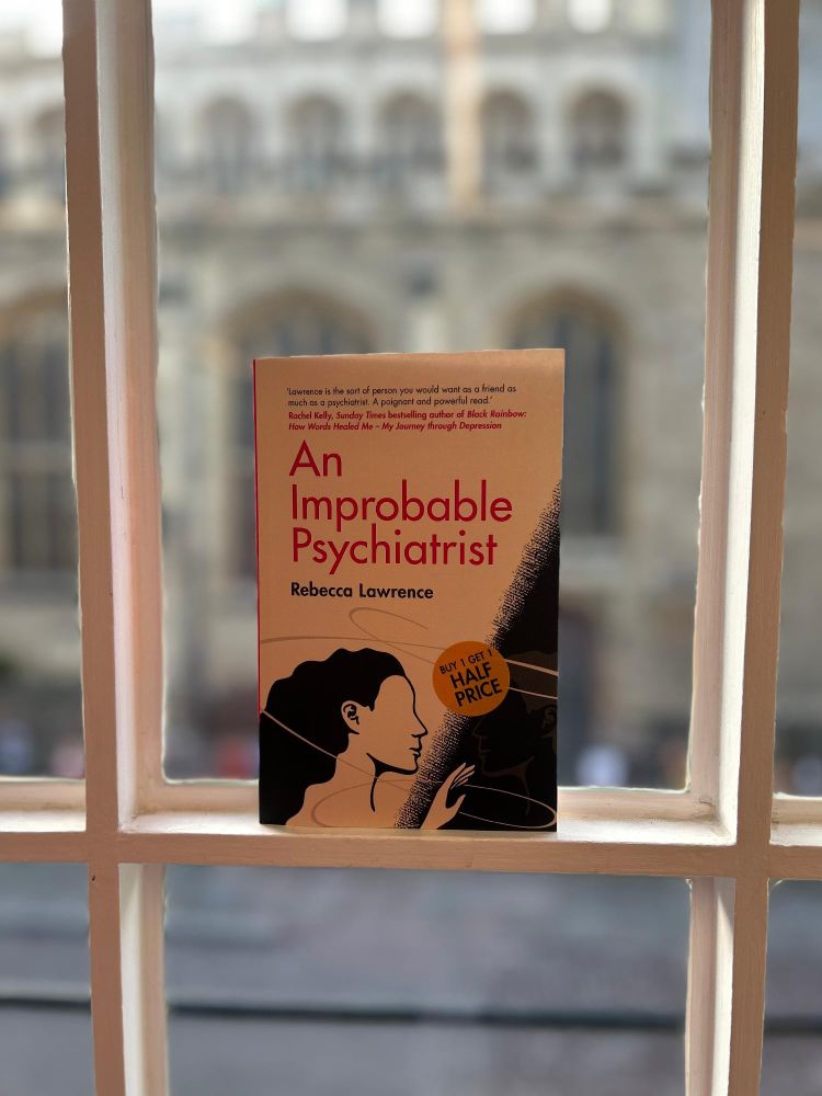 "An Improbable Psychiatrist" book by Rebecca Lawrence, displayed on a windowsill with a blurred background of a church.