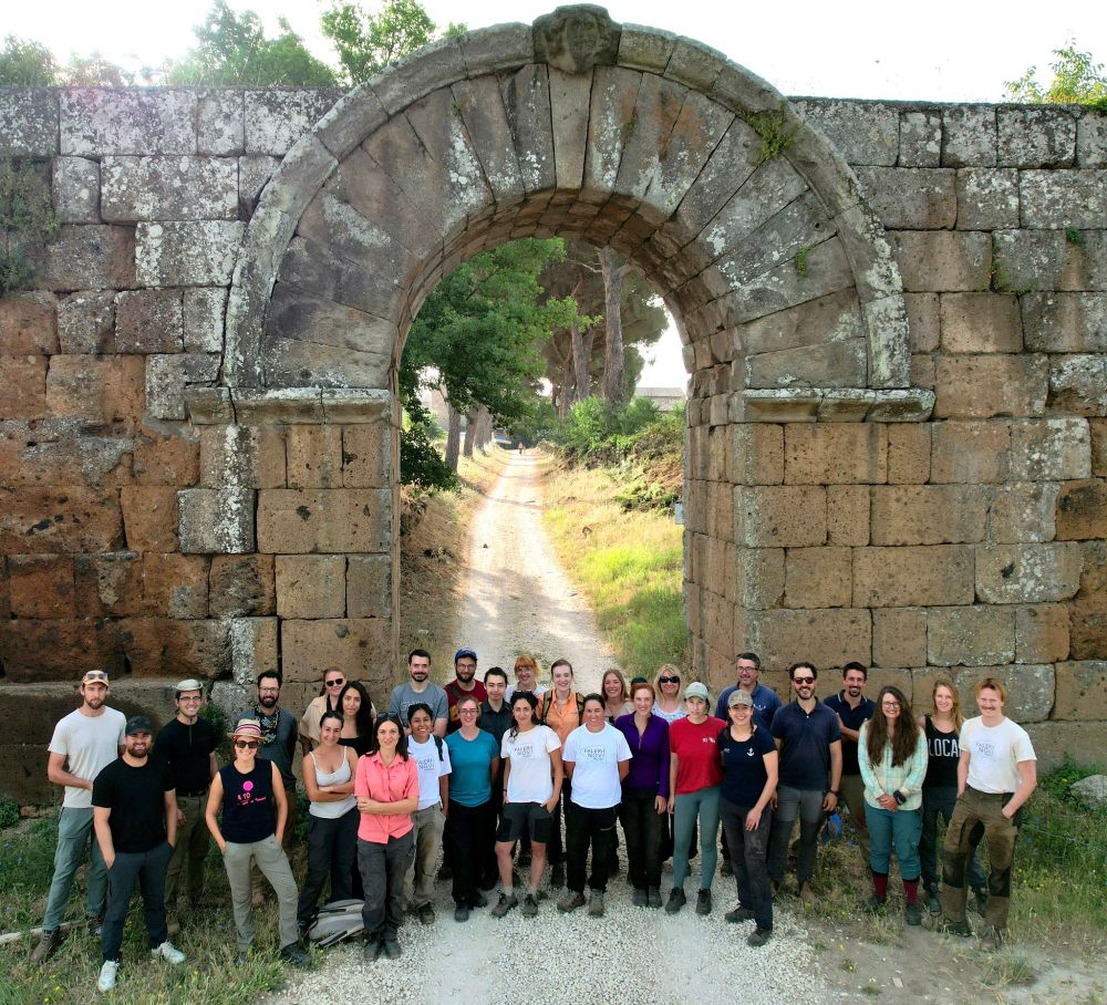Tall stone built arched gateway with an unpaved road stretching beyond it through a sunlit Mediterranean landscape. Standing in front are about thirty people in casual clothes and workboots.
