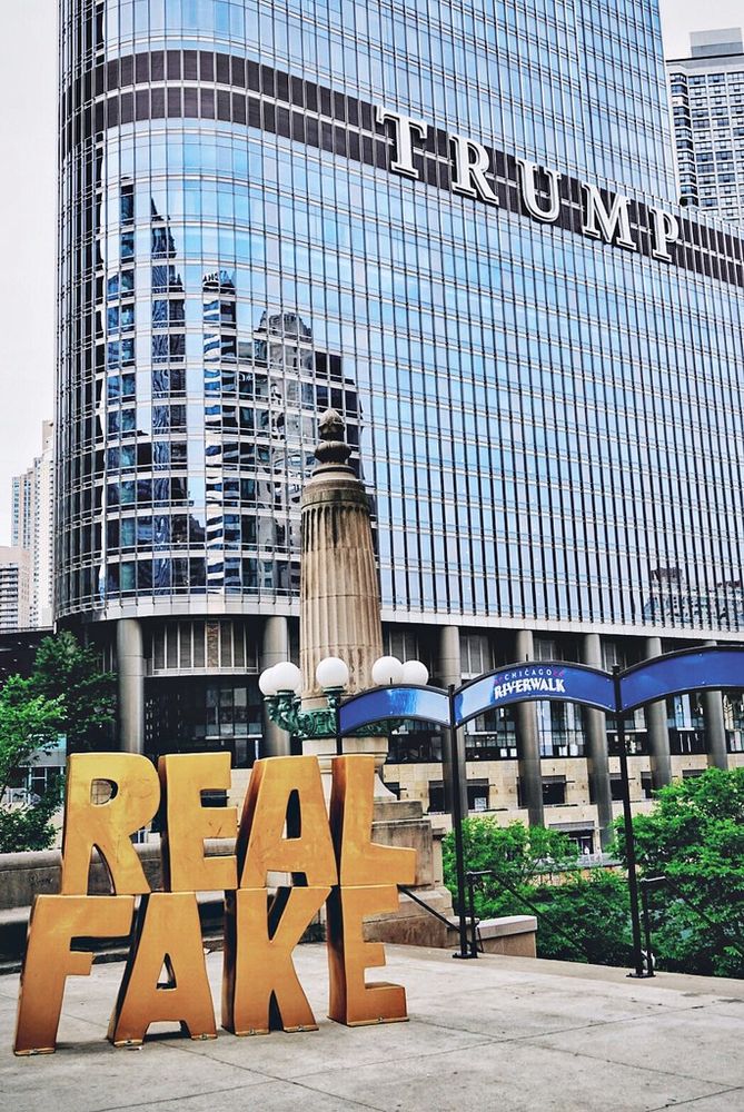 A large installation with the words "REAL FAKE" displayed in gold letters in front of the Trump International Hotel and Tower. The building's glass facade reflects nearby structures, and there are green trees and a sign for the Chicago Riverwalk.