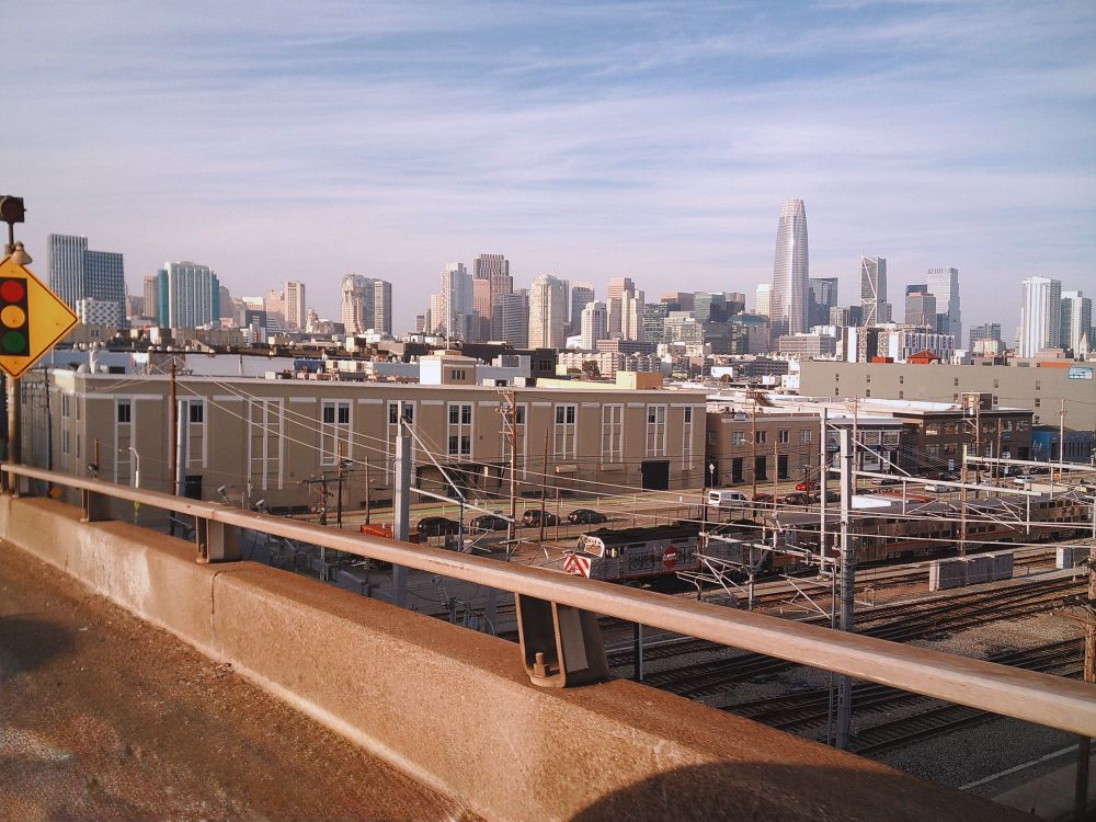 A city skyline features modern buildings, including a prominent tall structure, viewed from an elevated area. In the foreground, there are railway tracks and industrial buildings, with traffic signals visible. The scene is set under a clear sky.