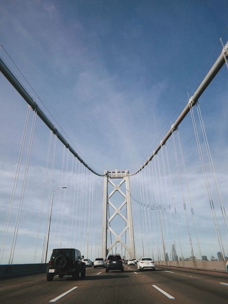 Photo of a bridge while driving on it. Incoming city skyline to the right. A few cars ahead.