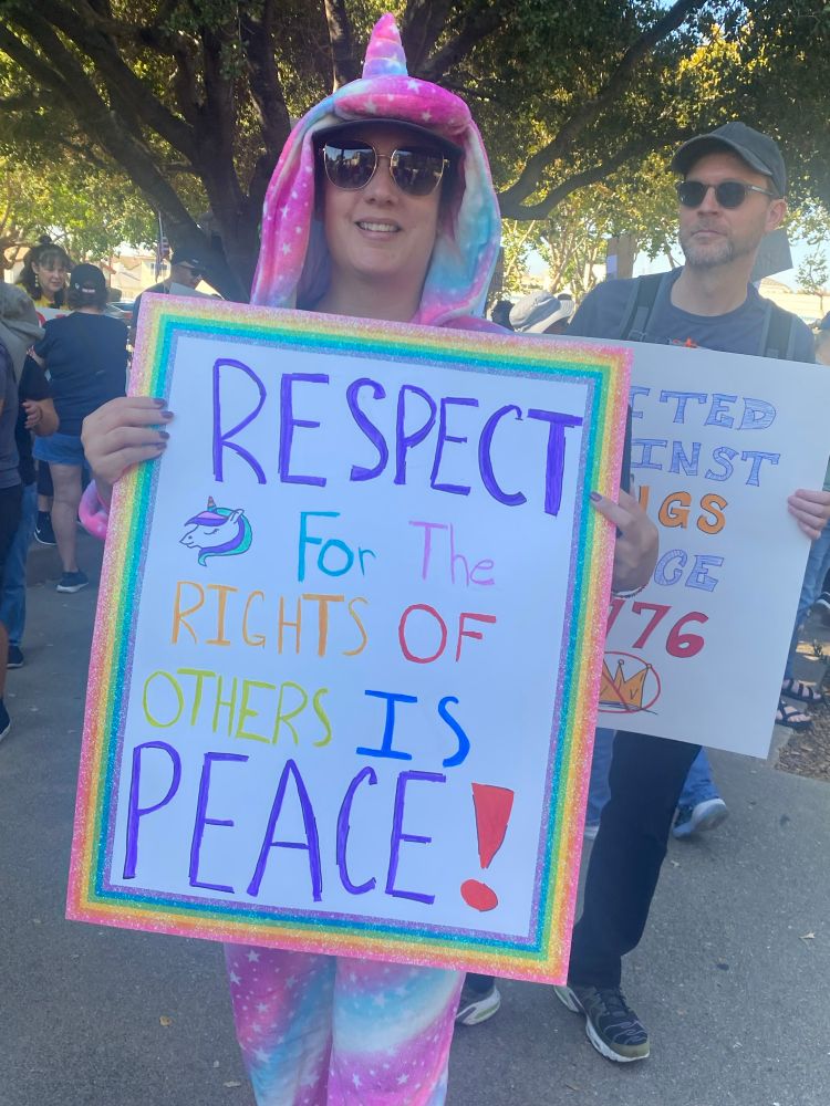 Person in a unicorn onesie with a sign that says “Respect for the rights of other is peace!”