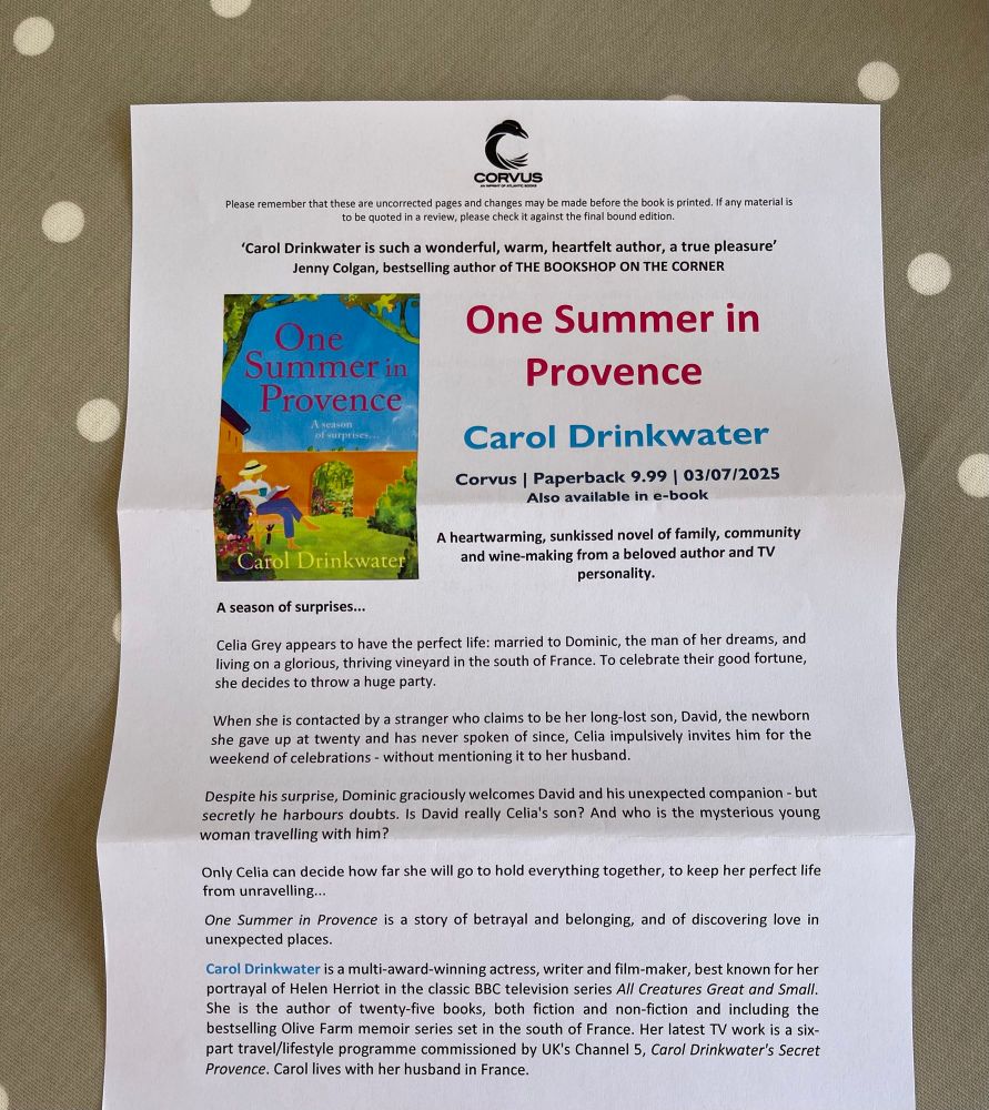 A printed promotional sheet for One Summer in Provence by Carol Drinkwater is placed on a polka-dotted tablecloth. The sheet includes the book cover, a summary of the story, and a brief biography of the author. The Corvus Books logo is visible at the top, along with a quote praising the author.