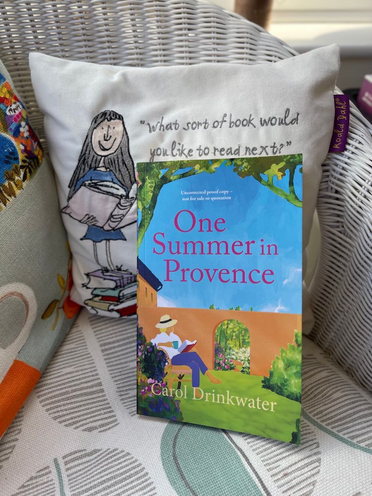 A proof copy of the book One Summer in Provence by Carol Drinkwater is propped up on a white wicker chair with a cushion behind it. The cushion features an illustration of a girl holding books and a quote that reads, “What sort of book would you like to read next?” The chair is part of a cozy reading nook with patterned cushions.