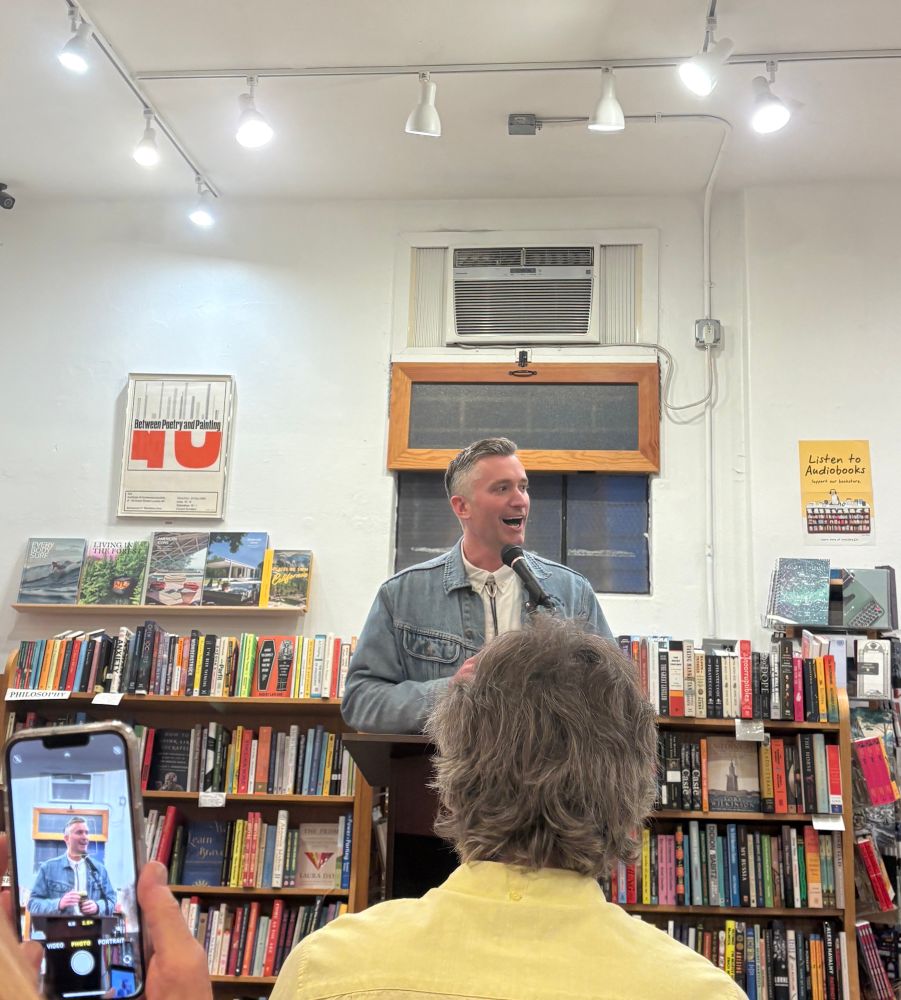 a man wearing jeans and a denim jacket over a white shirt standing at a podium reading an excerpt from his new novel for the Small Press Nite literary reading at the Book Catapult